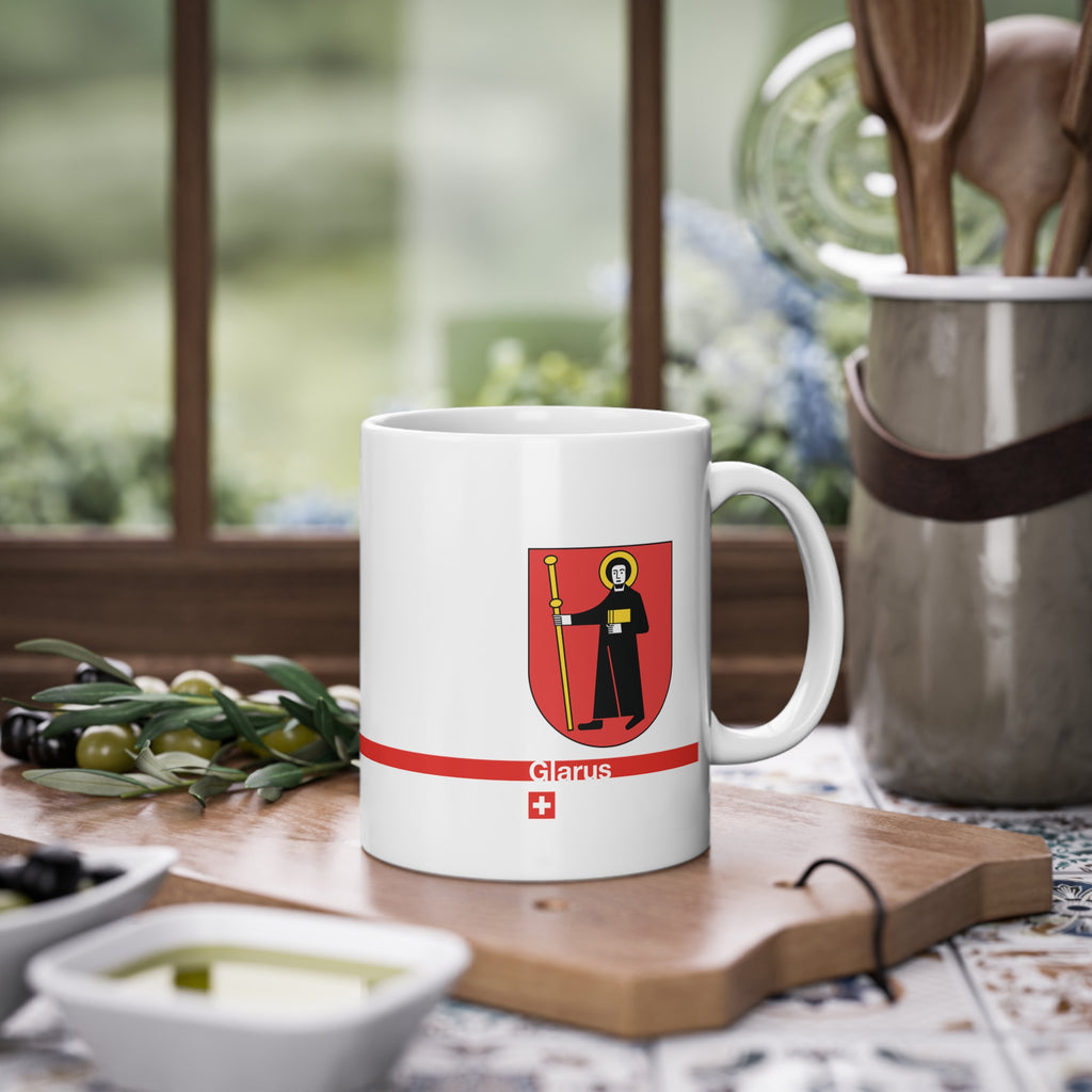 White ceramic mug with the red Canton of Glarus coat of arms featuring Saint Fridolin, a modern red line with 'Glarus' text and a small Swiss cross.